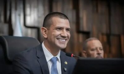 Homem de terno azul e gravata sorri em ambiente formal com microfone