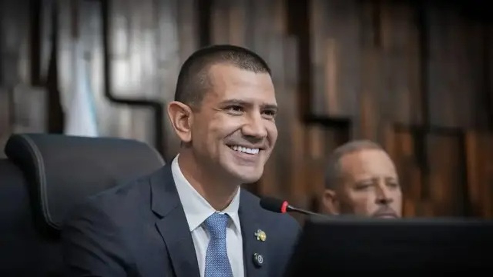 Homem de terno azul e gravata sorri em ambiente formal com microfone