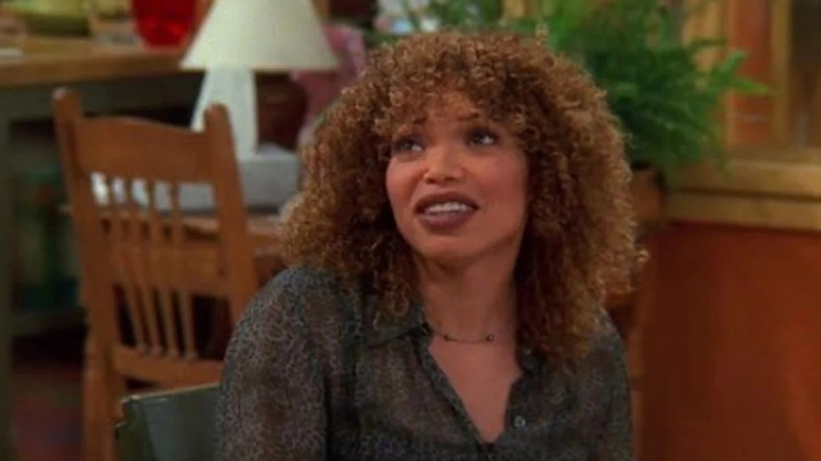 Tisha Campbell com cabelo cacheado, blusa estampada, sorrindo e olhando para o lado.