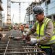 Trabalhador da construção civil com capacete e colete, operando ferramenta em vergalhões de aço