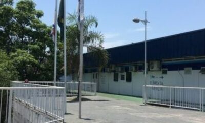 Fachada de clínica com letreiro "CLÍNICA DA FAMÍLIA", cercas brancas e postes com bandeiras
