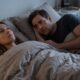 Casal, mulher e homem, deitados na cama sob cobertas, com expressões pensativas