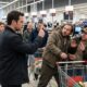 Um homem aponta para dois homens com expressões surpresas em uma fila de supermercado com carrinhos de compras