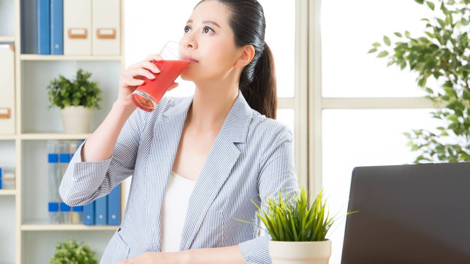 Mulher asiática bebendo suco vermelho de um copo de vidro em um escritório