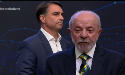 Lula em primeiro plano falando, Flávio Bolsonaro atrás, em debate com hashtag #DebateNaBand
