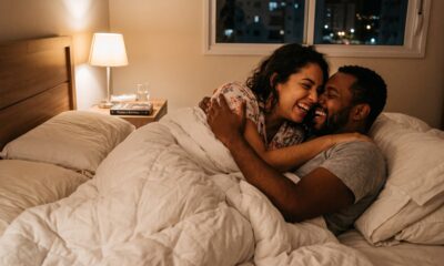 Casal sorridente e abraçado na cama, deitados sob um edredom branco, à noite em um quarto