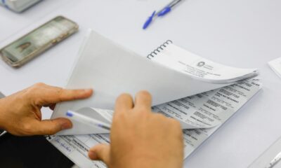 Mãos manuseiam caderno de votação com o cabeçalho 'Tribunal Regional Eleitoral Rio de Janeiro'. Smartphone desfocado ao fundo