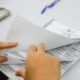 Mãos manuseiam caderno de votação com o cabeçalho 'Tribunal Regional Eleitoral Rio de Janeiro'. Smartphone desfocado ao fundo