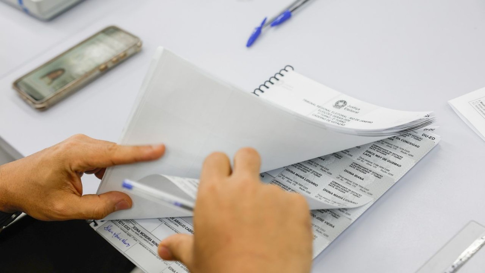 Mãos manuseiam caderno de votação com o cabeçalho 'Tribunal Regional Eleitoral Rio de Janeiro'. Smartphone desfocado ao fundo