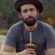 Matteus Amaral com chapéu, camiseta cinza "Alegrete Matteus", barba, segurando chimarrão em área rural