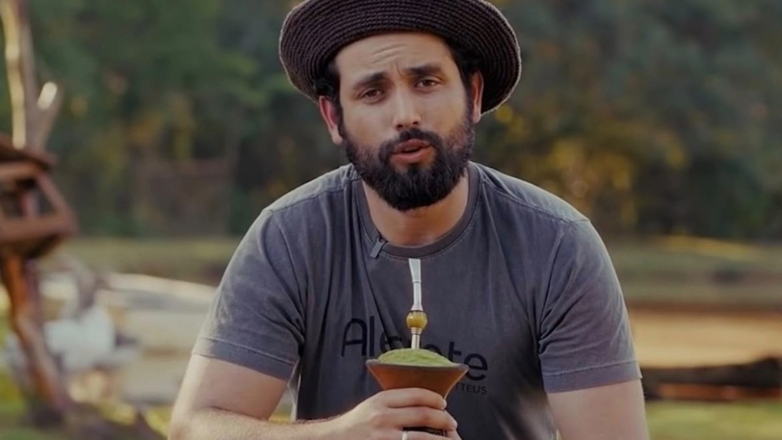 Matteus Amaral com chapéu, camiseta cinza "Alegrete Matteus", barba, segurando chimarrão em área rural