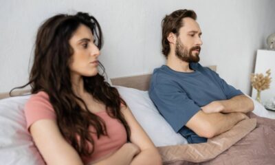 Casal de costas na cama, braços cruzados, com semblantes sérios, indicando tensão ou distância