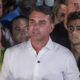 Flávio Bolsonaro de camisa branca fala, cercado por apoiadores. Homem à esquerda veste camisa CBF com 'Flávio Bolsonaro 22'