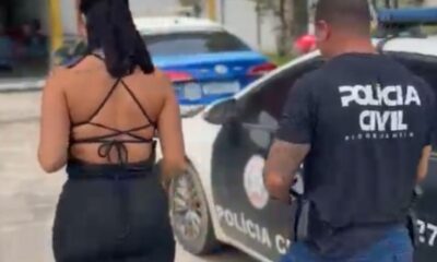 Mulher de costas em roupa preta ao lado de policial civil e viatura da polícia