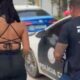 Mulher de costas em roupa preta ao lado de policial civil e viatura da polícia