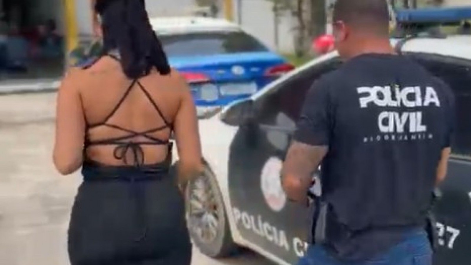 Mulher de costas em roupa preta ao lado de policial civil e viatura da polícia