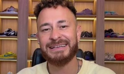 Homem de cabelo encaracolado, barba e piercing no nariz sorrindo, com estante de tênis ao fundo