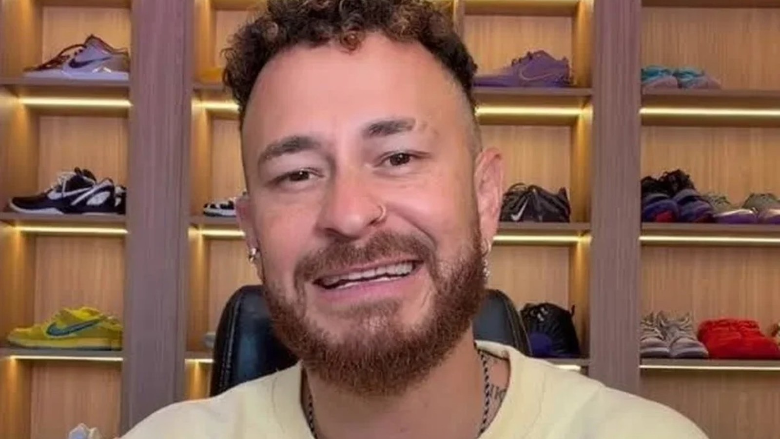 Homem de cabelo encaracolado, barba e piercing no nariz sorrindo, com estante de tênis ao fundo