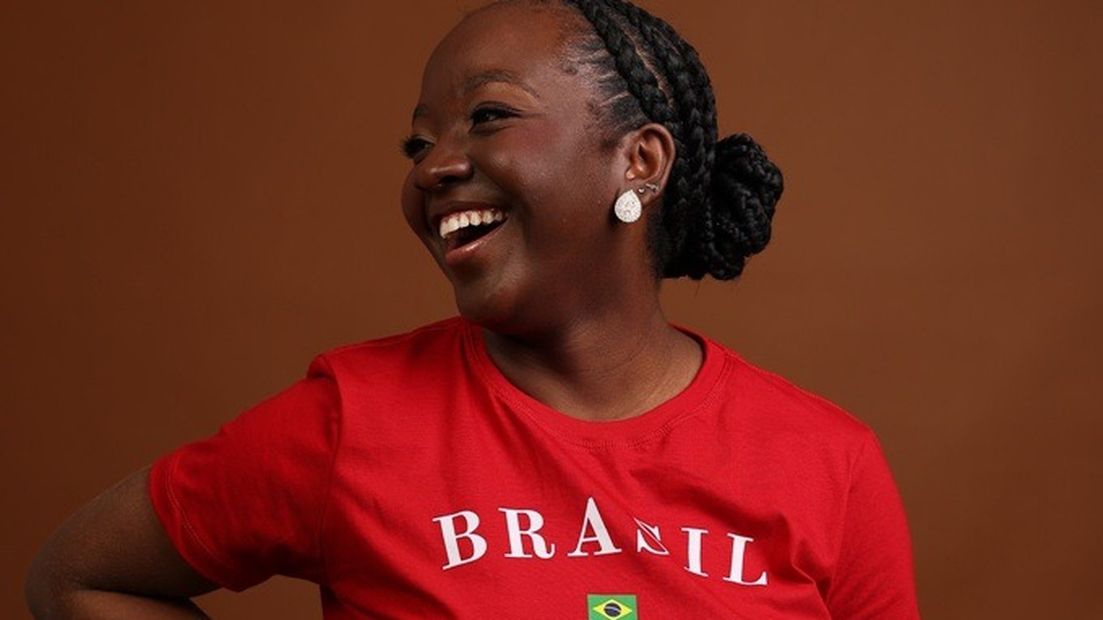 Mulher de pele escura sorrindo, cabelo trançado, camiseta vermelha com "BRASIL" e bandeira