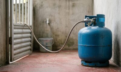 Botijão de gás azul conectado por mangueira e regulador, em piso vermelho com parede cinza