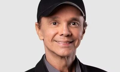 Cantor Netinho sorrindo, com boné preto, jaqueta escura e camisa cinza