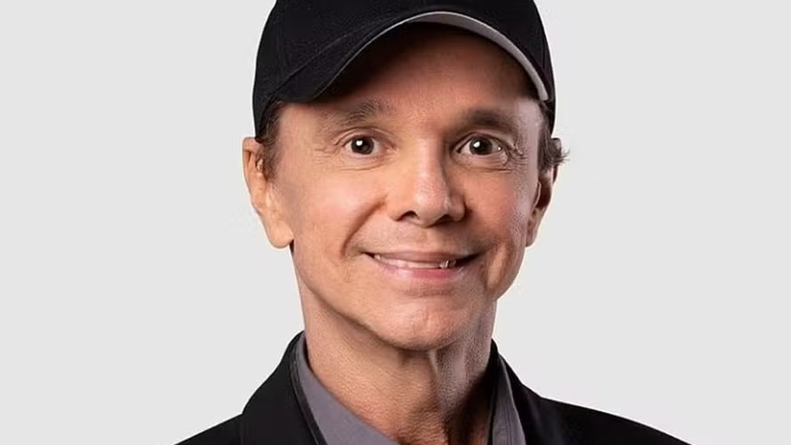Cantor Netinho sorrindo, com boné preto, jaqueta escura e camisa cinza
