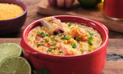 Canjiquinha com costelinha suína em tigela vermelha, guarnecida com cebolinha. Limões e milho ao fundo