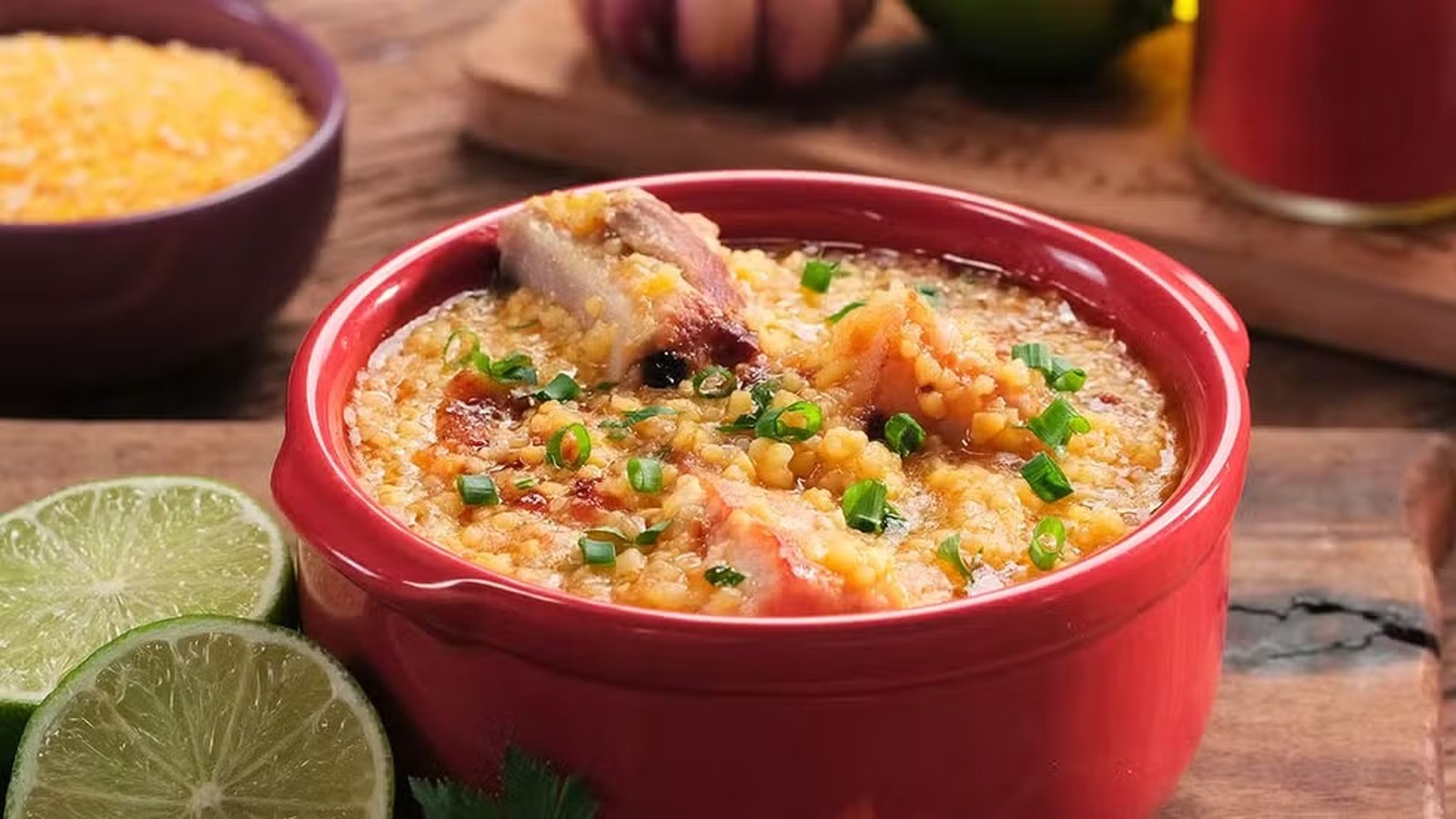 Canjiquinha com costelinha suína em tigela vermelha, guarnecida com cebolinha. Limões e milho ao fundo