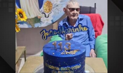 Homem de óculos e camisa azul com bolo azul escrito '113' e 'João Marinho'