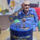 Homem de óculos e camisa azul com bolo azul escrito '113' e 'João Marinho'