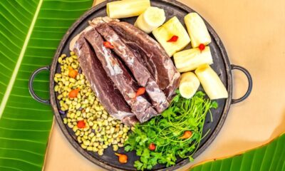 Carne de sol, mandioca, feijão, coentro e pimentas em uma panela rústica sobre folha de bananeira