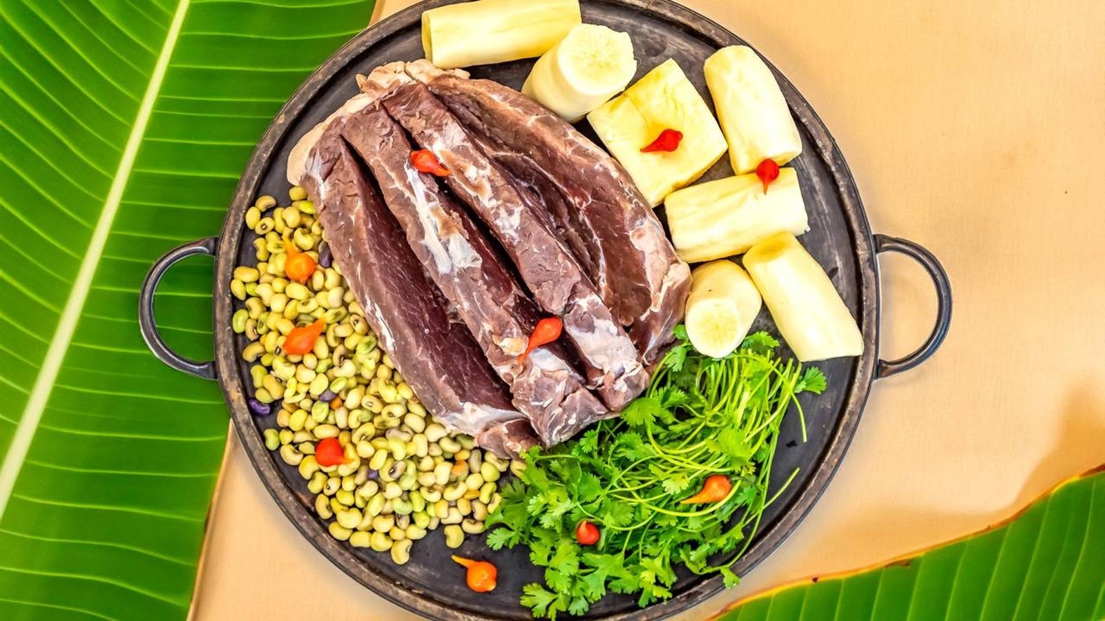 Carne de sol, mandioca, feijão, coentro e pimentas em uma panela rústica sobre folha de bananeira