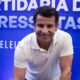 Homem sorridente em camisa polo branca, com fundo azul e texto "PROGRESSISTAS ELEI"