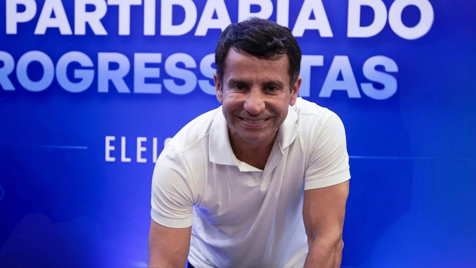 Homem sorridente em camisa polo branca, com fundo azul e texto "PROGRESSISTAS ELEI"