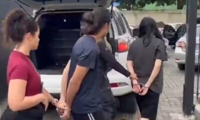 Duas mulheres algemadas sendo escoltadas por uma terceira, próximo a um SUV branco.