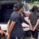 Duas mulheres algemadas sendo escoltadas por uma terceira, próximo a um SUV branco.