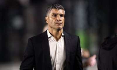 Homem de meia-idade com cabelo e barba grisalhos, blazer preto e camisa creme, olhar sério