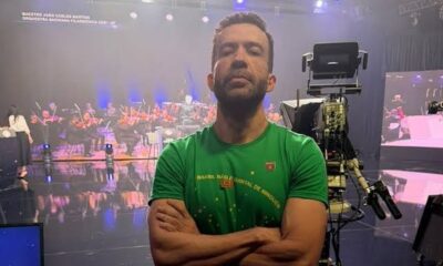Homem de barba em camisa verde, braços cruzados, em estúdio de TV com orquestra, tela de fundo