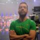 Homem de barba em camisa verde, braços cruzados, em estúdio de TV com orquestra, tela de fundo
