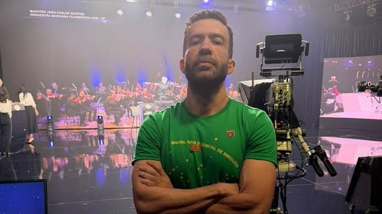 Homem de barba em camisa verde, braços cruzados, em estúdio de TV com orquestra, tela de fundo