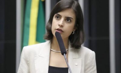 Mulher jovem de blazer claro fala em microfone em ambiente formal, bandeira verde e amarela