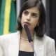 Mulher jovem de blazer claro fala em microfone em ambiente formal, bandeira verde e amarela