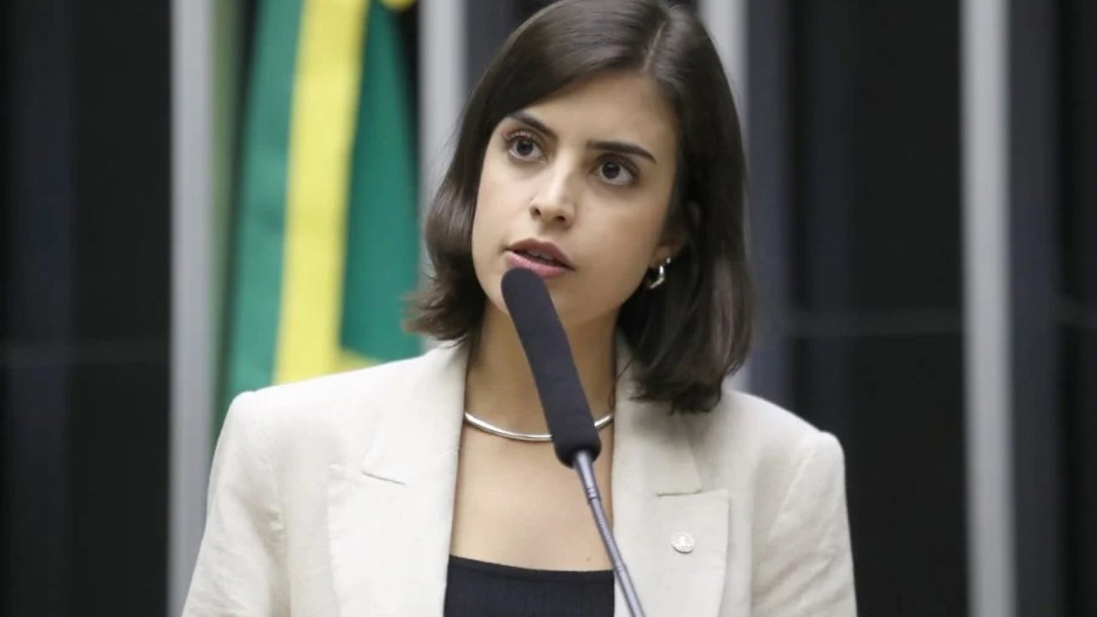 Mulher jovem de blazer claro fala em microfone em ambiente formal, bandeira verde e amarela