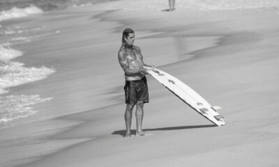 Homem sem camisa com cabelo molhado e tatuagens segurando prancha de surfe na praia