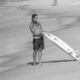Homem sem camisa com cabelo molhado e tatuagens segurando prancha de surfe na praia