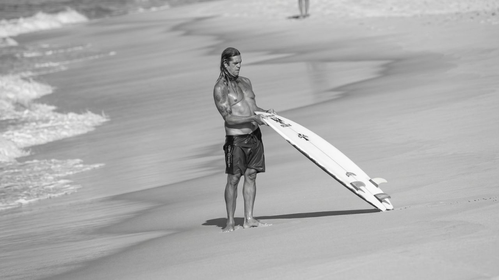 Homem sem camisa com cabelo molhado e tatuagens segurando prancha de surfe na praia
