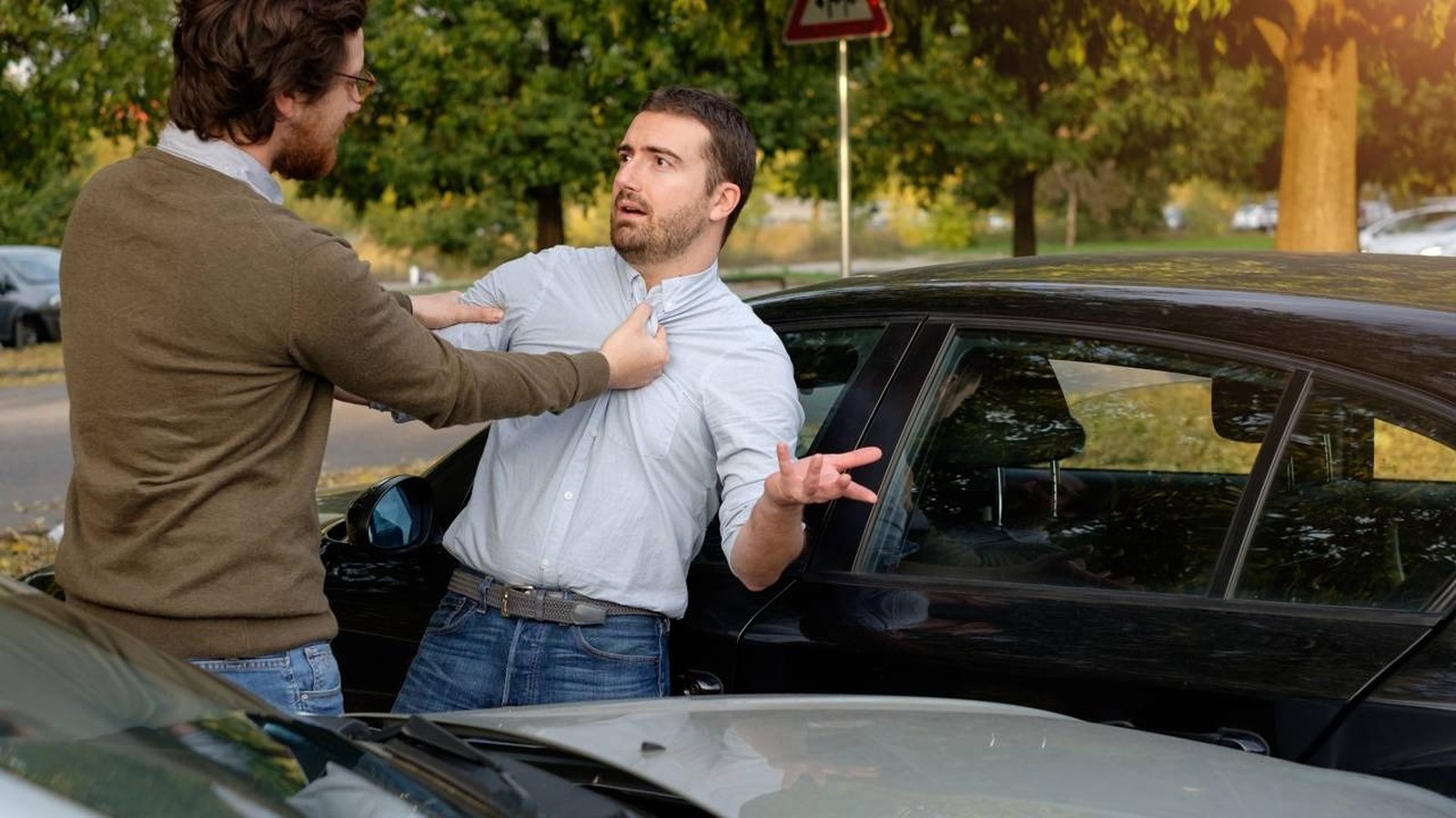 Dois homens brigam, um segura o outro pela camisa, perto de carros na rua