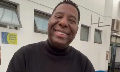 Netinho de Paula sorrindo. Camisa preta. Fundo com janelas e placa 'AQUI TEM TELEFONE PÚBLICO'