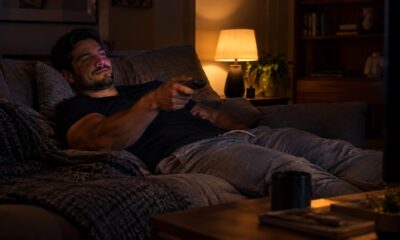 Homem deitado no sofá em sala escura, sorrindo e segurando um controle remoto na mão