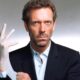 Hugh Laurie sério, vestindo luva médica, camisa azul e terno preto, contra fundo cinza
